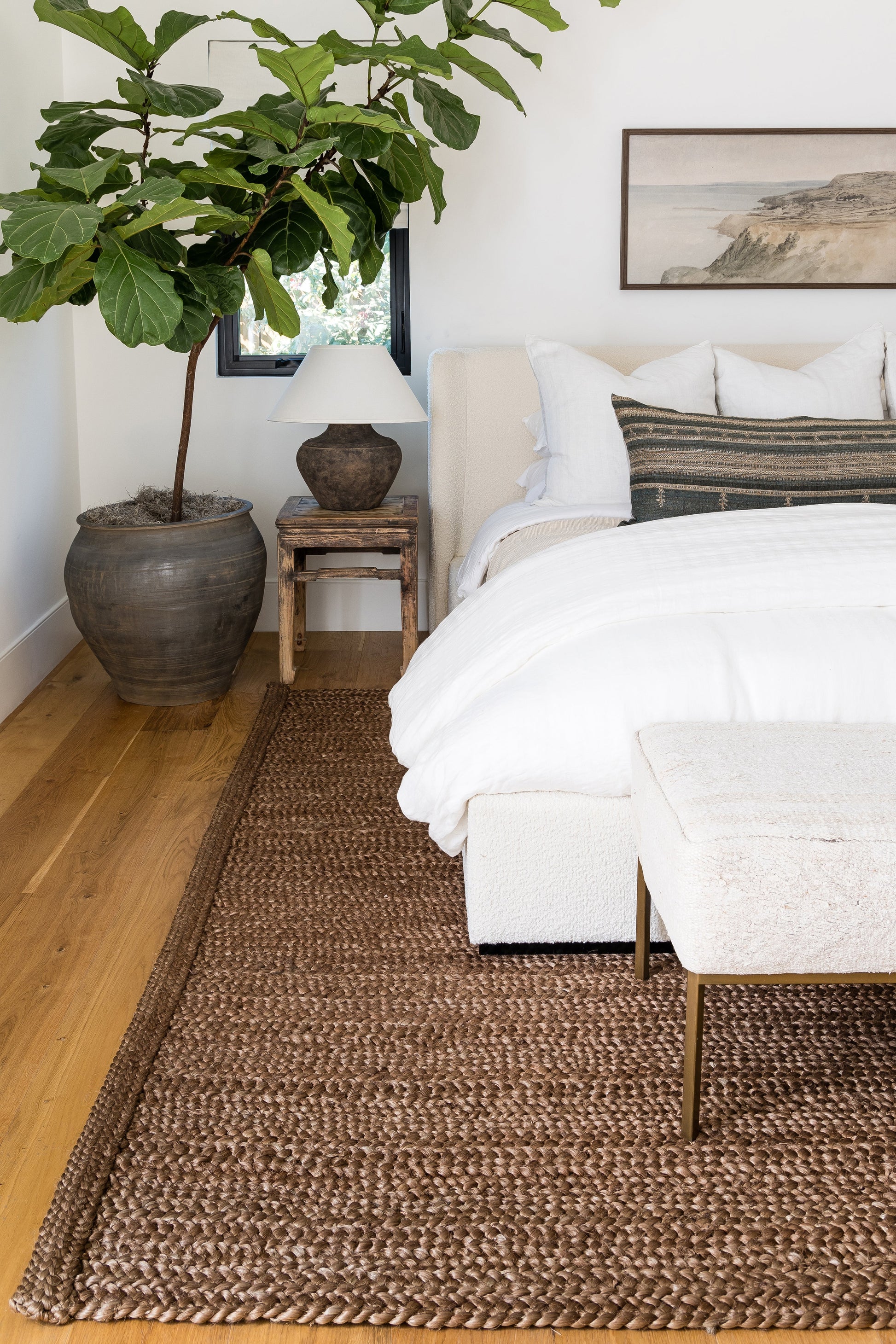 Bright bedroom with white bedding, beige upholstered bed, fiddle-leaf fig in a ceramic pot, rustic nightstand and jute rug.