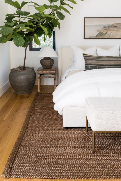 Large fiddle-leaf fig in a ceramic pot, white bedding, jute rug, wooden side table & lamp, beige bench.
