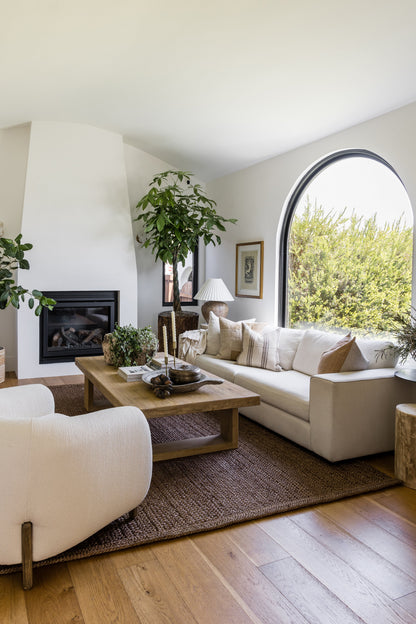 Bright living room with a round 6' rug, cream upholstered sofa & armchairs, wooden coffee table, arched window, fireplace, greenery & neutral decor.