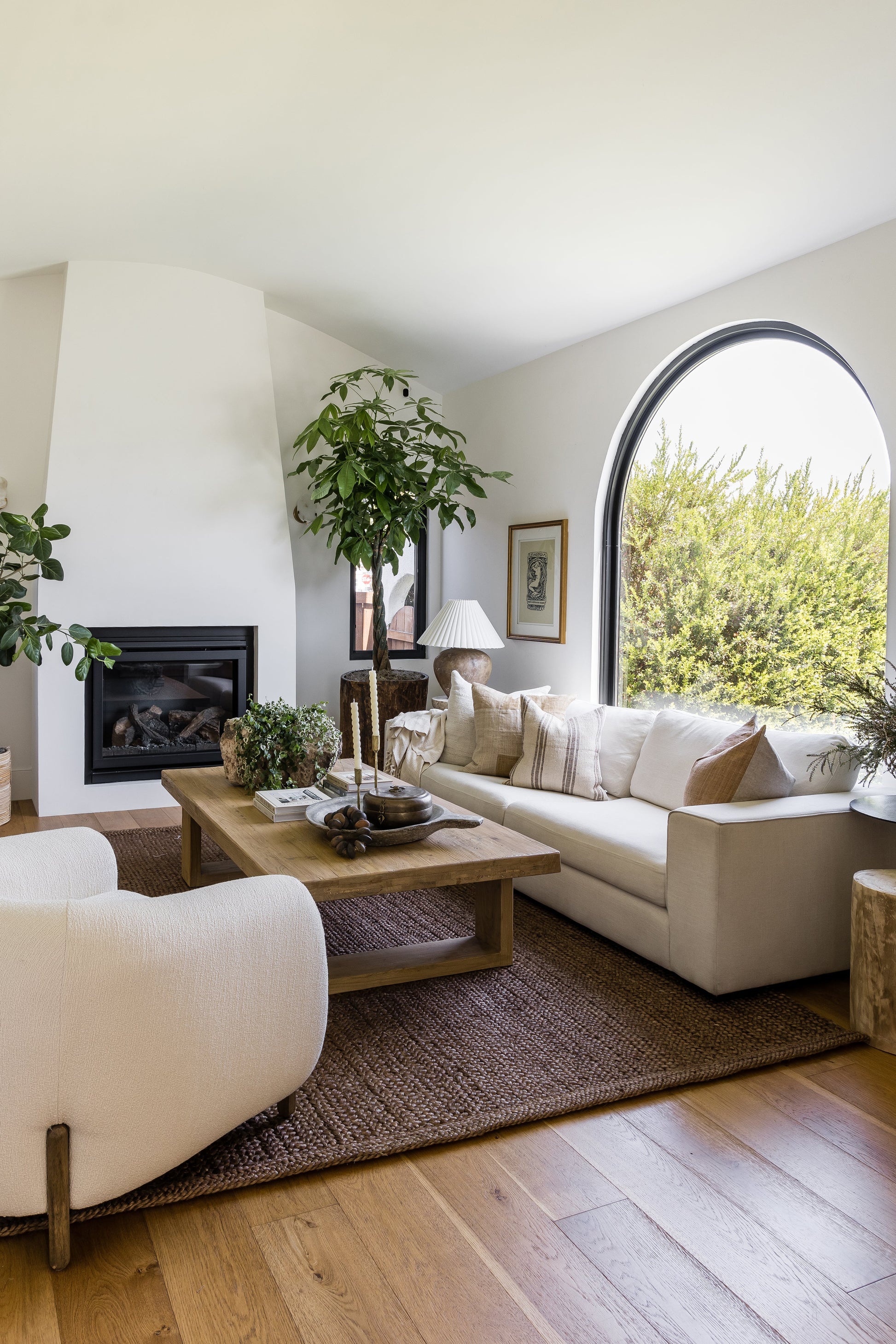 Bright modern living room with cream sofa, curved armchair, wooden coffee table, fireplace, arched window, and plants.