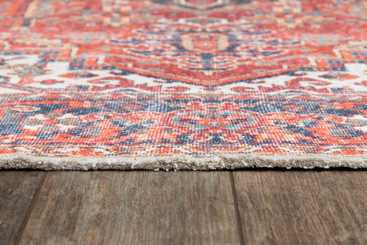 Close-up of a vibrant red-orange patterned rug with blue and cream accents, fringe edge on a dark wood floor.