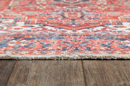 Close-up of a vibrant red oriental rug with orange and blue patterns and fringe along the edge on a wooden floor.