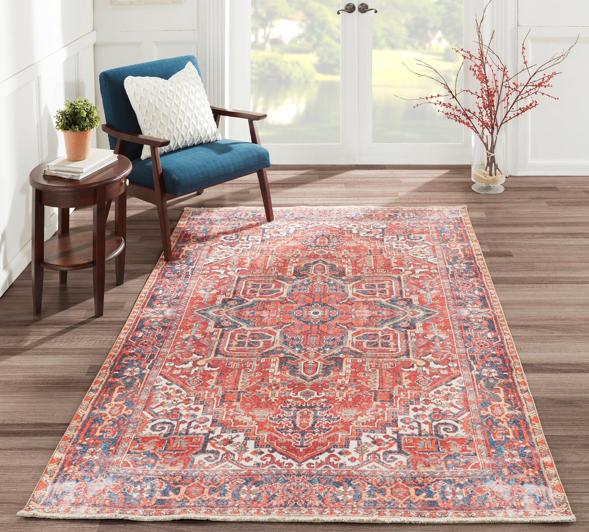 Intricate red and blue Oriental rug on wood floor beside a blue upholstered armchair, round wood side table, plant, and vase with branches.