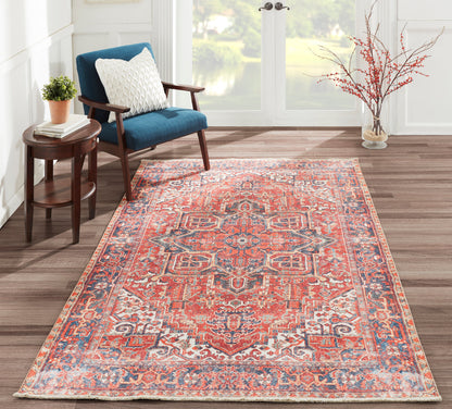 Intricate red and blue Oriental rug on wood floor beside a blue upholstered armchair, round wood side table, plant, and vase with branches.