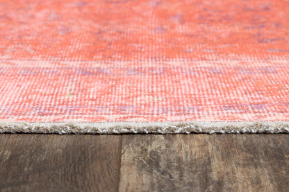 Coral pink woven rug with lavender accents and a fringed edge on wood floor.