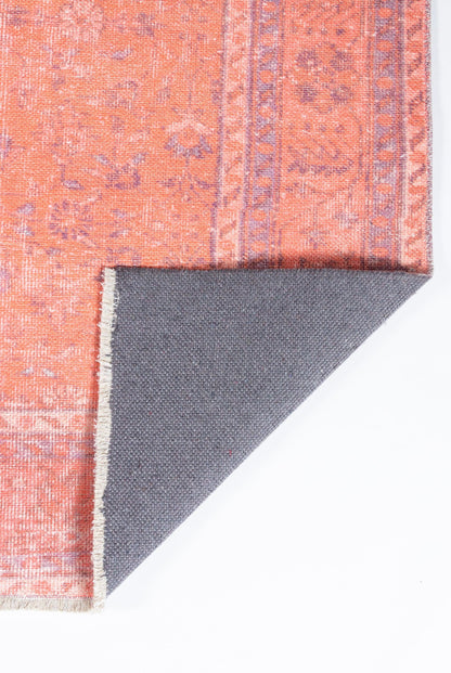 Terracotta orange vintage-style rug with subtle pink floral patterns and bordered motif; folded corner reveals grey backing.