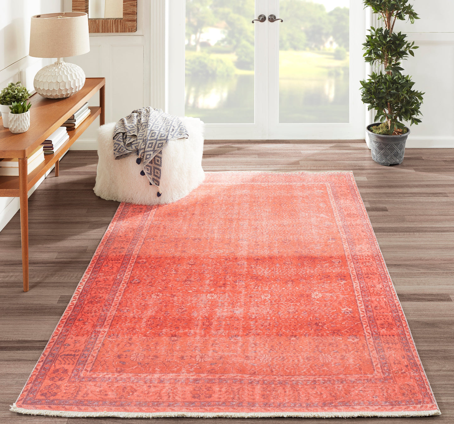 Coral rug with faded pattern in a bright living room; white fluffy pouf, gray geometric throw, wooden console and plant.