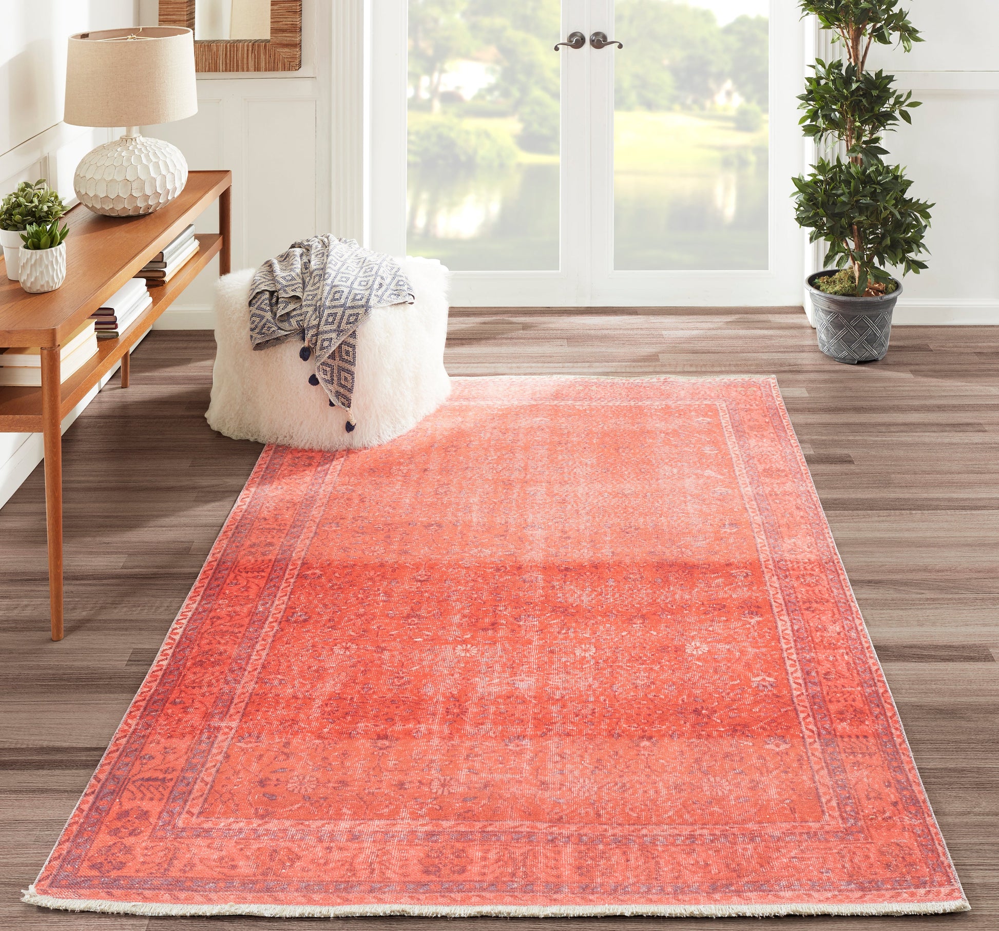 Coral rug with faded pattern in a bright living room; white fluffy pouf, gray geometric throw, wooden console and plant.