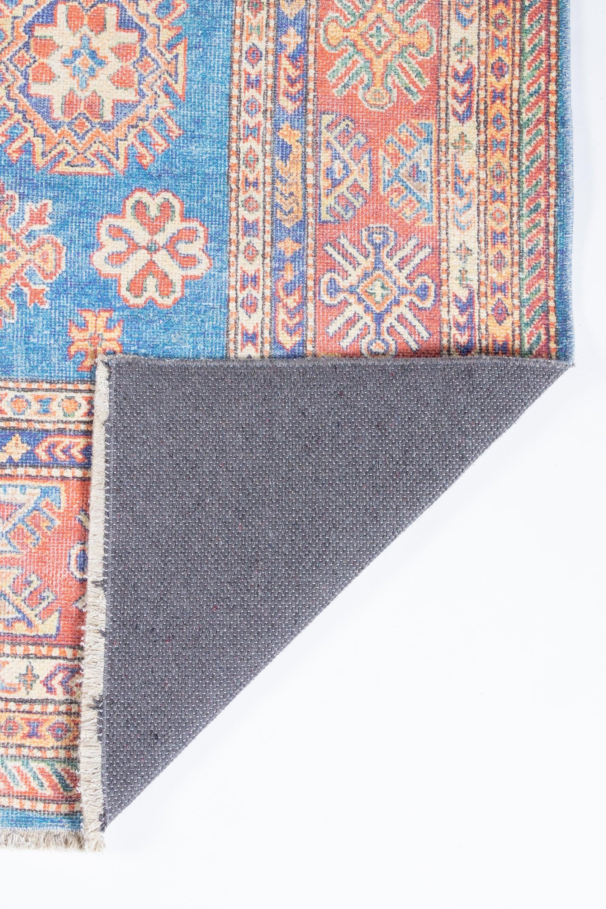 Blue and coral oriental rug with geometric medallions, intricate border, woven fringe; folded corner reveals gray backing.