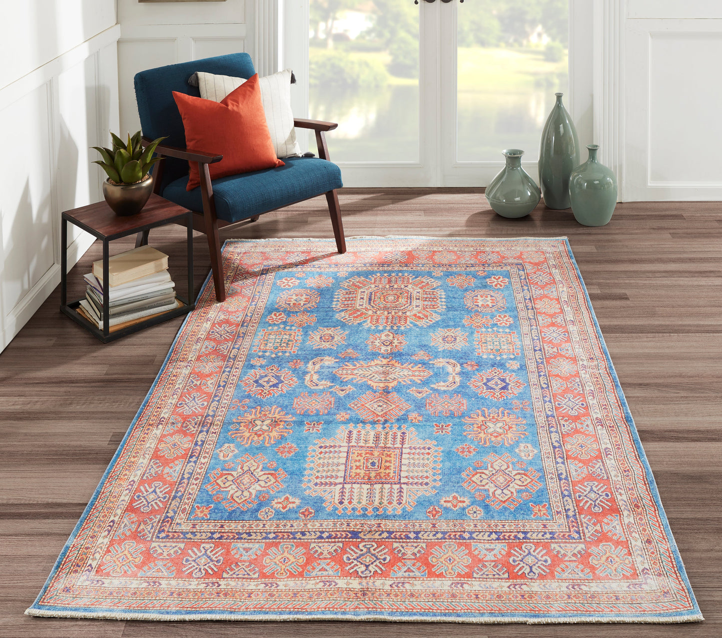 Blue and coral oriental-style area rug with geometric medallions on a wood floor beside a blue velvet chair.
