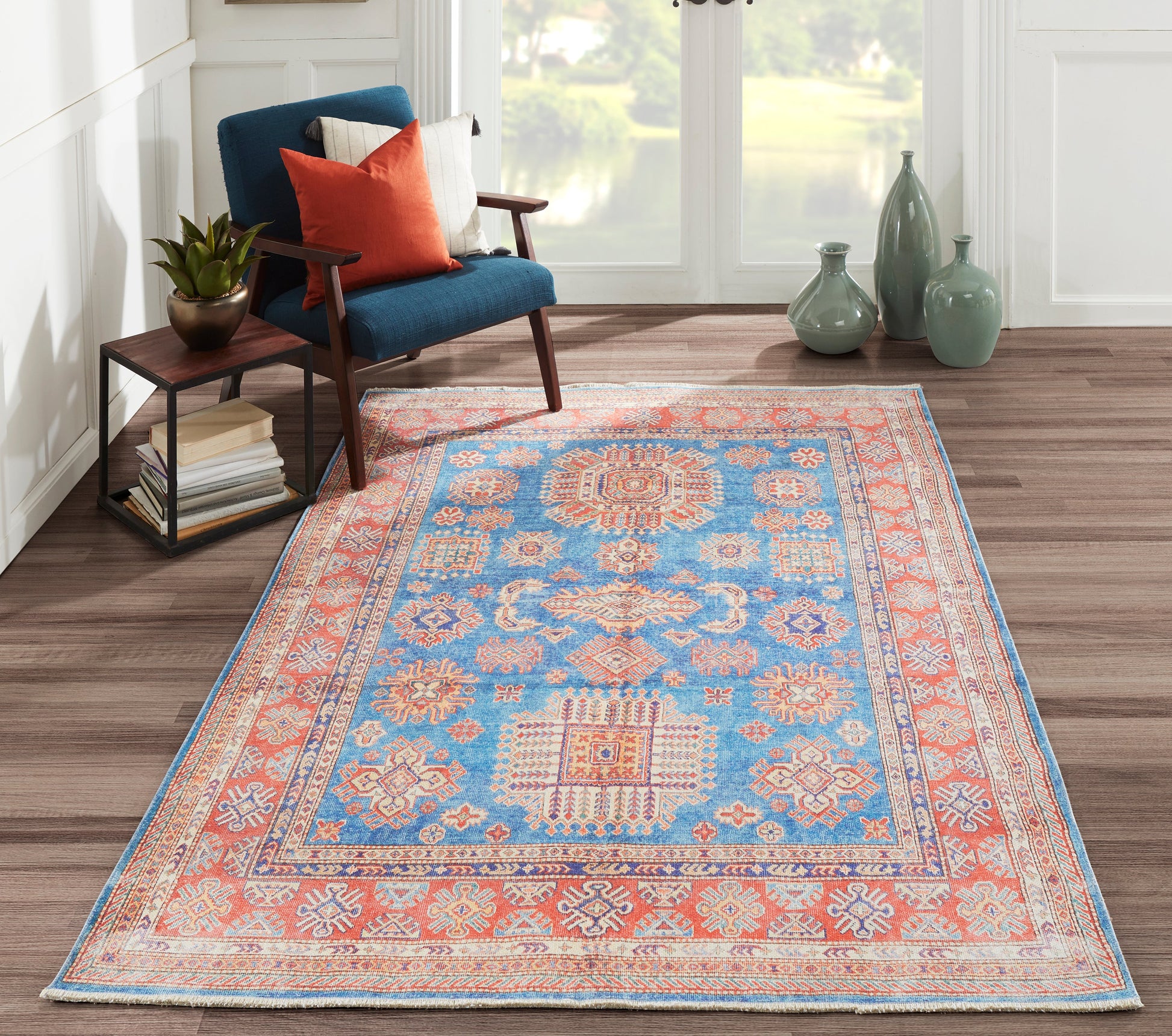 Blue oriental rug with orange border on wood floor, blue armchair with orange cushion, side table, and green vases by window.