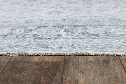 Gray damask-pattern rug with white fringe edge on dark wood floor.