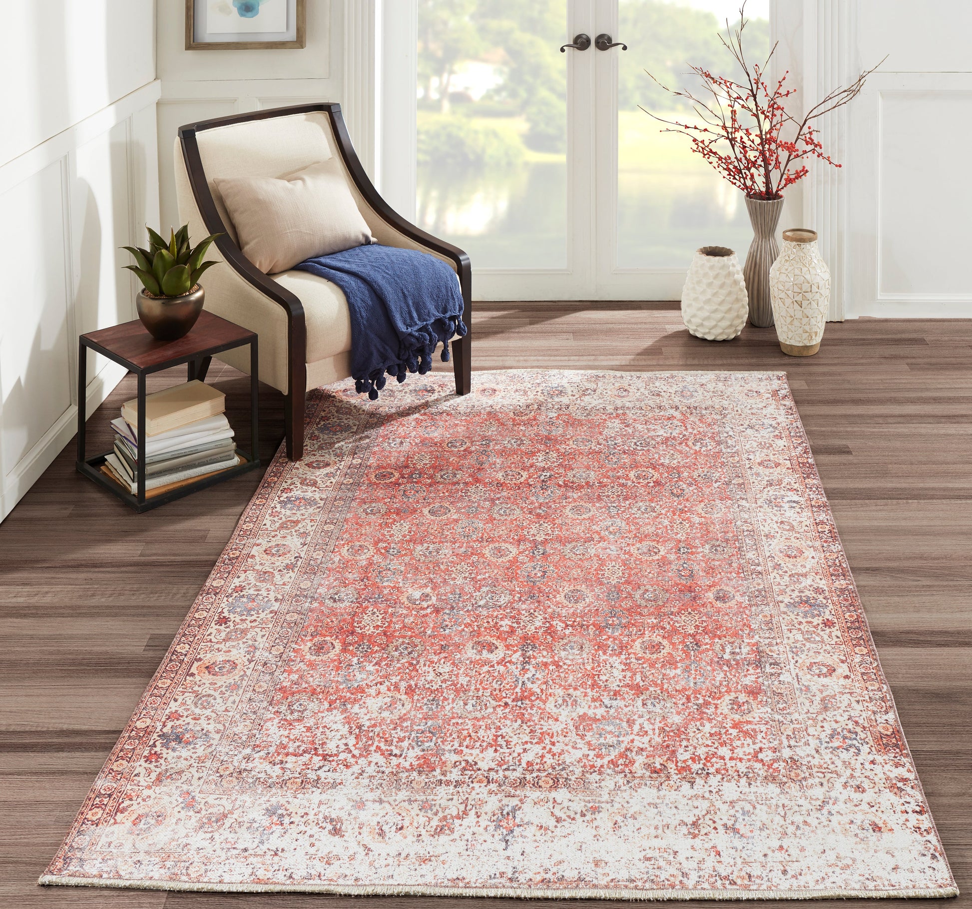 Red-toned Persian-style rug in a bright living room with beige upholstered chair, navy throw, side table & plant.