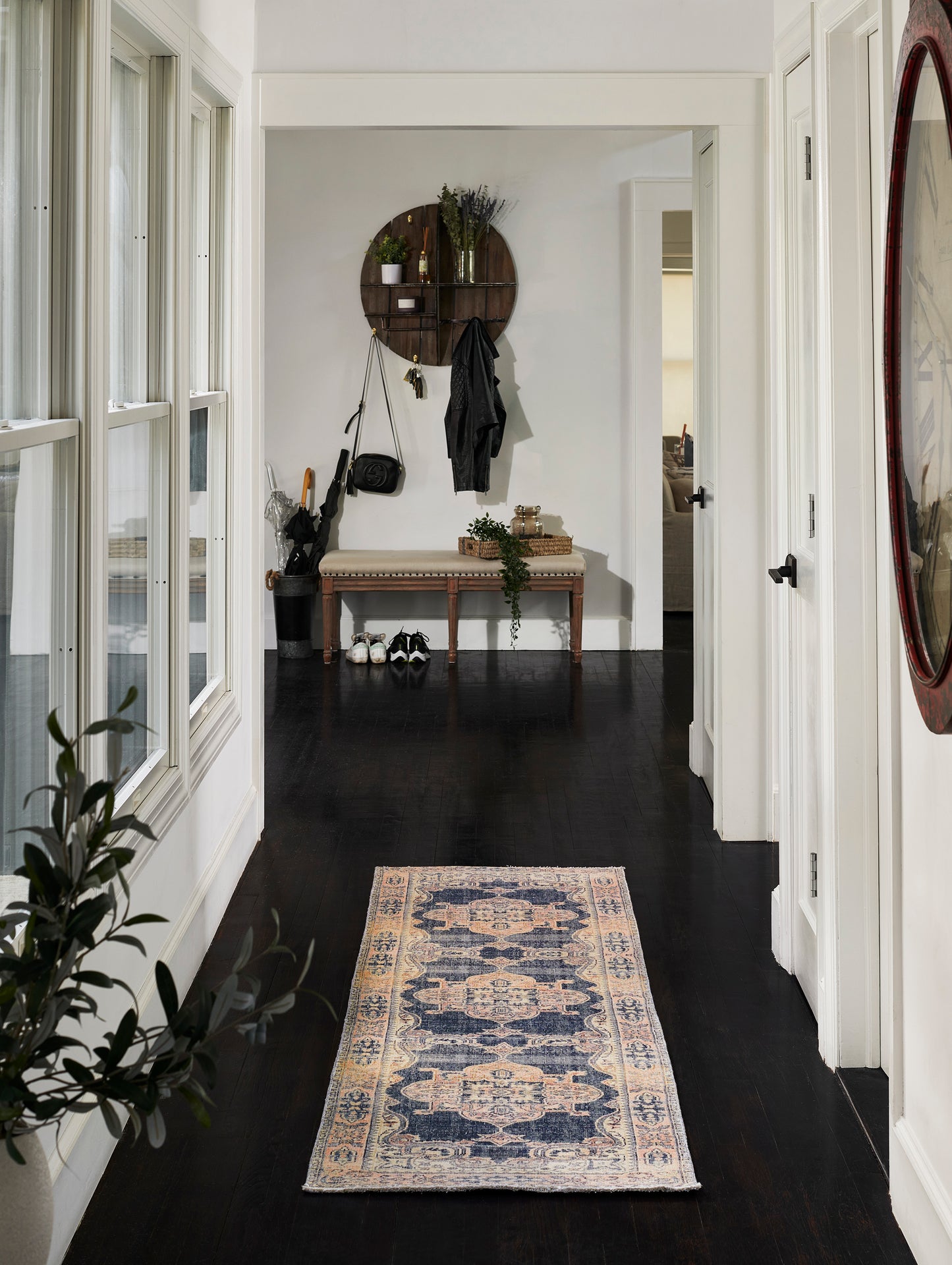Narrow entry hall with dark wood floor, cream-blue patterned rug, round wall shelf with plants, bench and coats.