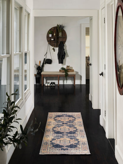 Narrow entry hall with dark wood floor, cream-blue patterned rug, round wall shelf with plants, bench and coats.