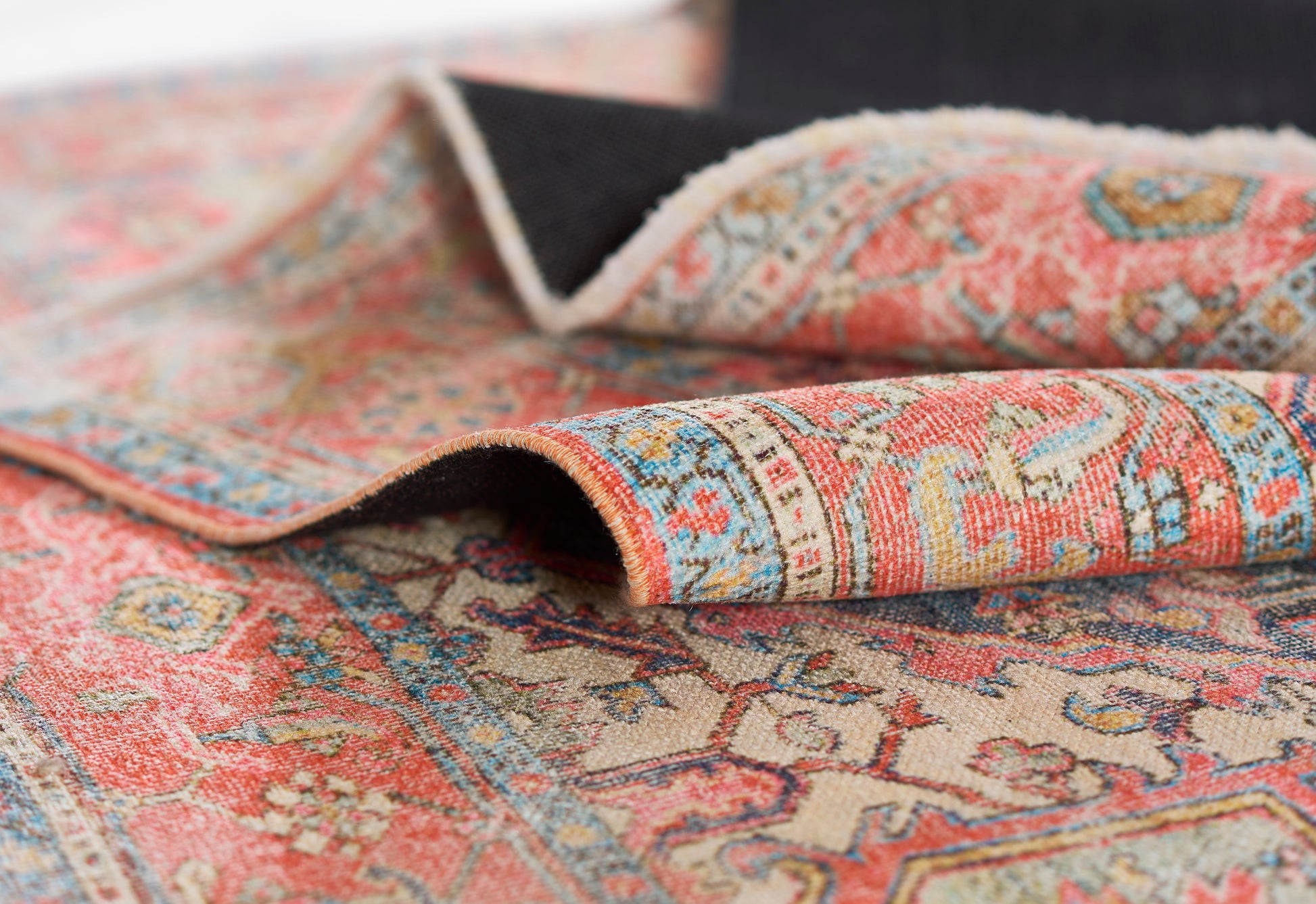 Close-up of ornate oriental rug in coral pink, blue and beige with floral motifs and folded corner.