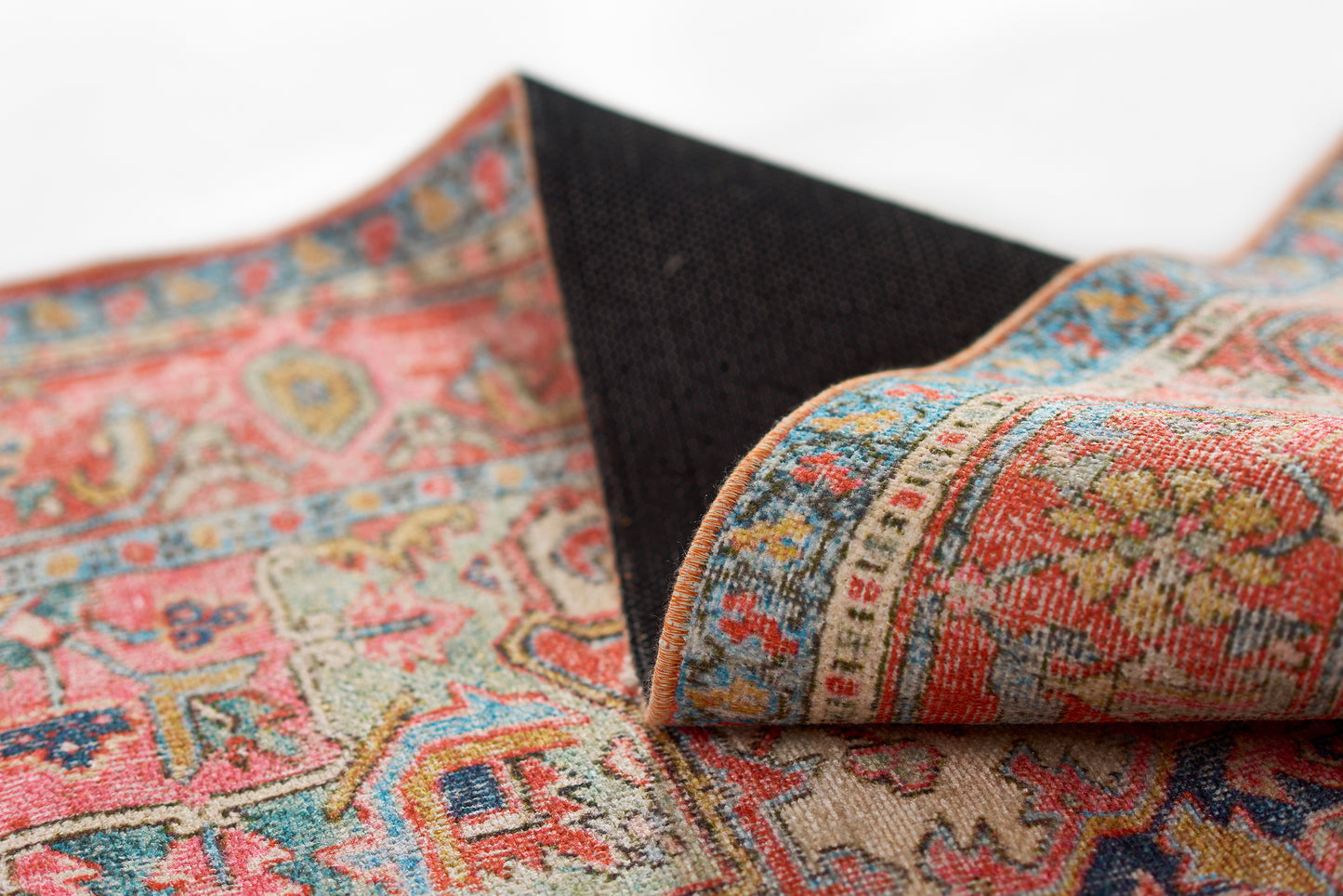 Close-up of colorful Persian-style rug with pink, teal, blue, and beige motifs; folded corner reveals navy underside.