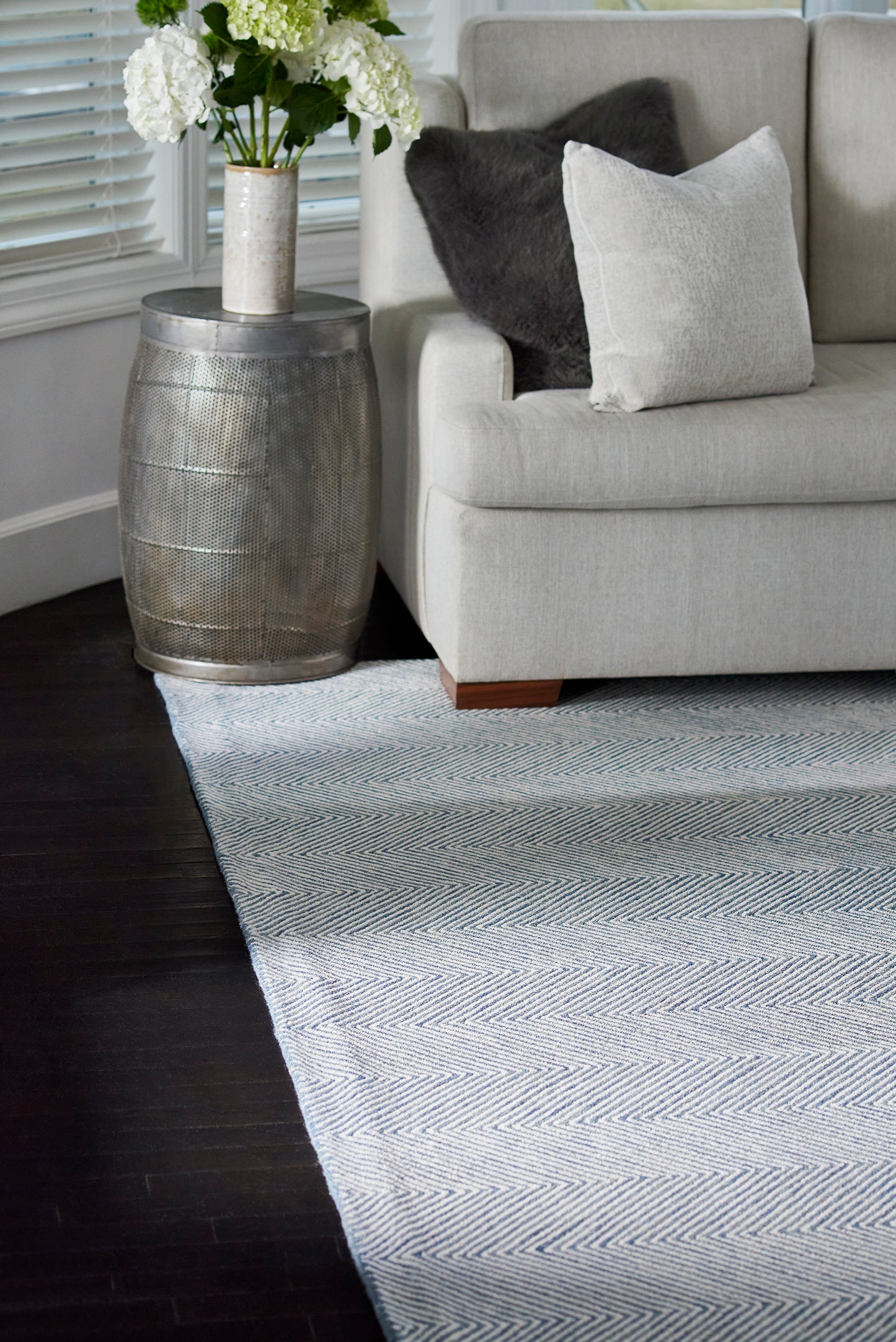 Light gray sofa with white pillow and dark throw, silver drum-side table with white hydrangeas, and a blue chevron rug