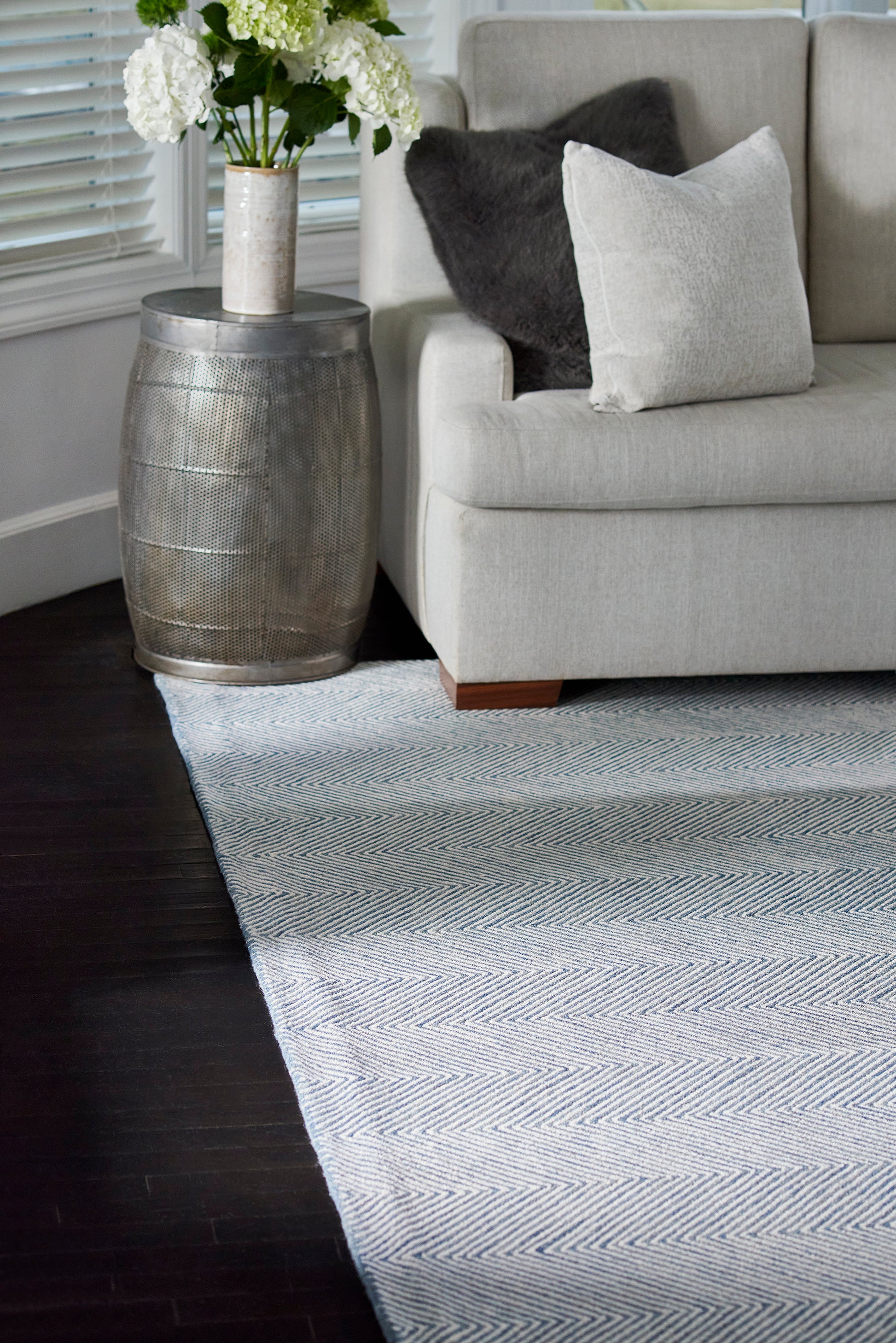 Light gray sofa with white pillow and dark throw, silver drum-side table with white hydrangeas, and a blue chevron rug