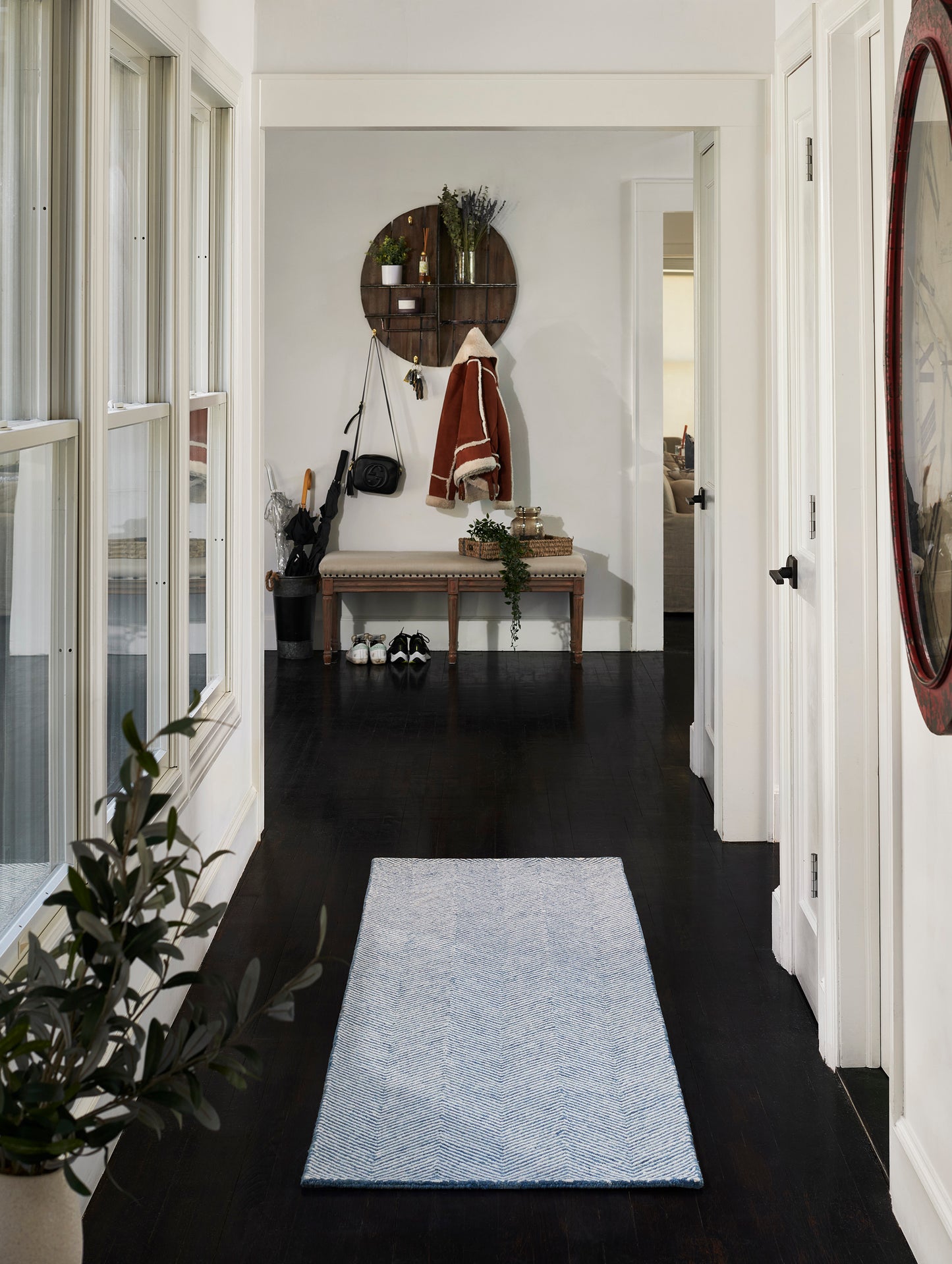 Blue chevron runner rug on dark hardwood hallway with white walls, potted plant, bench, coat rack, and round wall shelf.