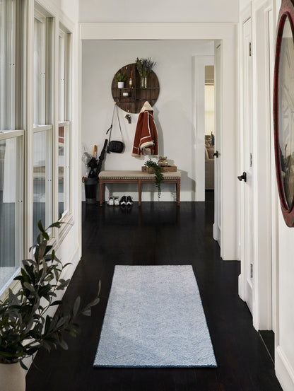 Blue chevron runner rug on dark hardwood hallway with white walls, potted plant, bench, coat rack, and round wall shelf.