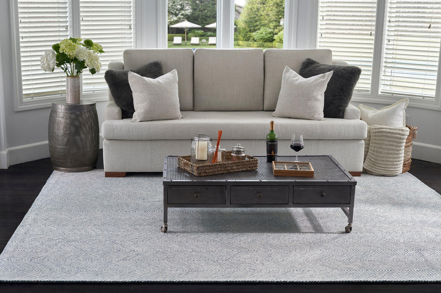 Light-gray sofa with white/gray pillows, metal 3-drawer coffee table on casters, wicker tray, wine bottle, hydrangea vase, blue-gray rug.