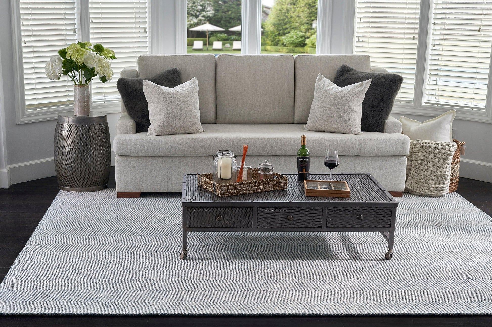 Light-gray sofa with white/gray pillows, metal 3-drawer coffee table on casters, wicker tray, wine bottle, hydrangea vase, blue-gray rug.