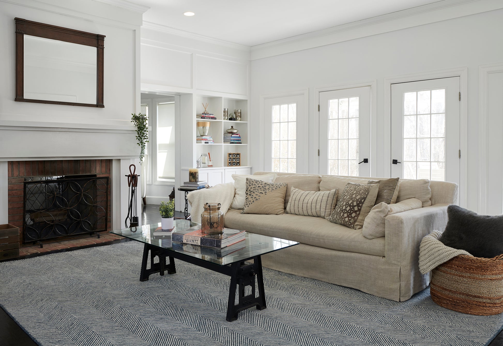Bright white living room with beige sofa, glass coffee table on striped rug, brick fireplace, shelves, and wicker basket.