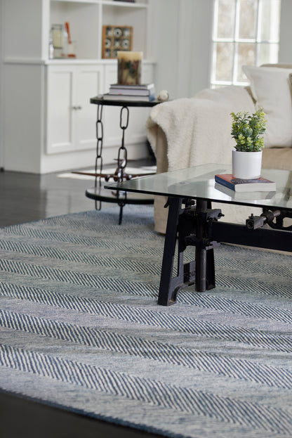 Living room scene: glass coffee table on blue-gray chevron rug, potted plant on books, cream throw on beige sofa, white shelving.