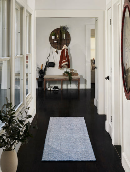 Hallway with dark wood floors, light blue chevron rug, upholstered bench, wood shelf, coats and bag, red-framed mirror.