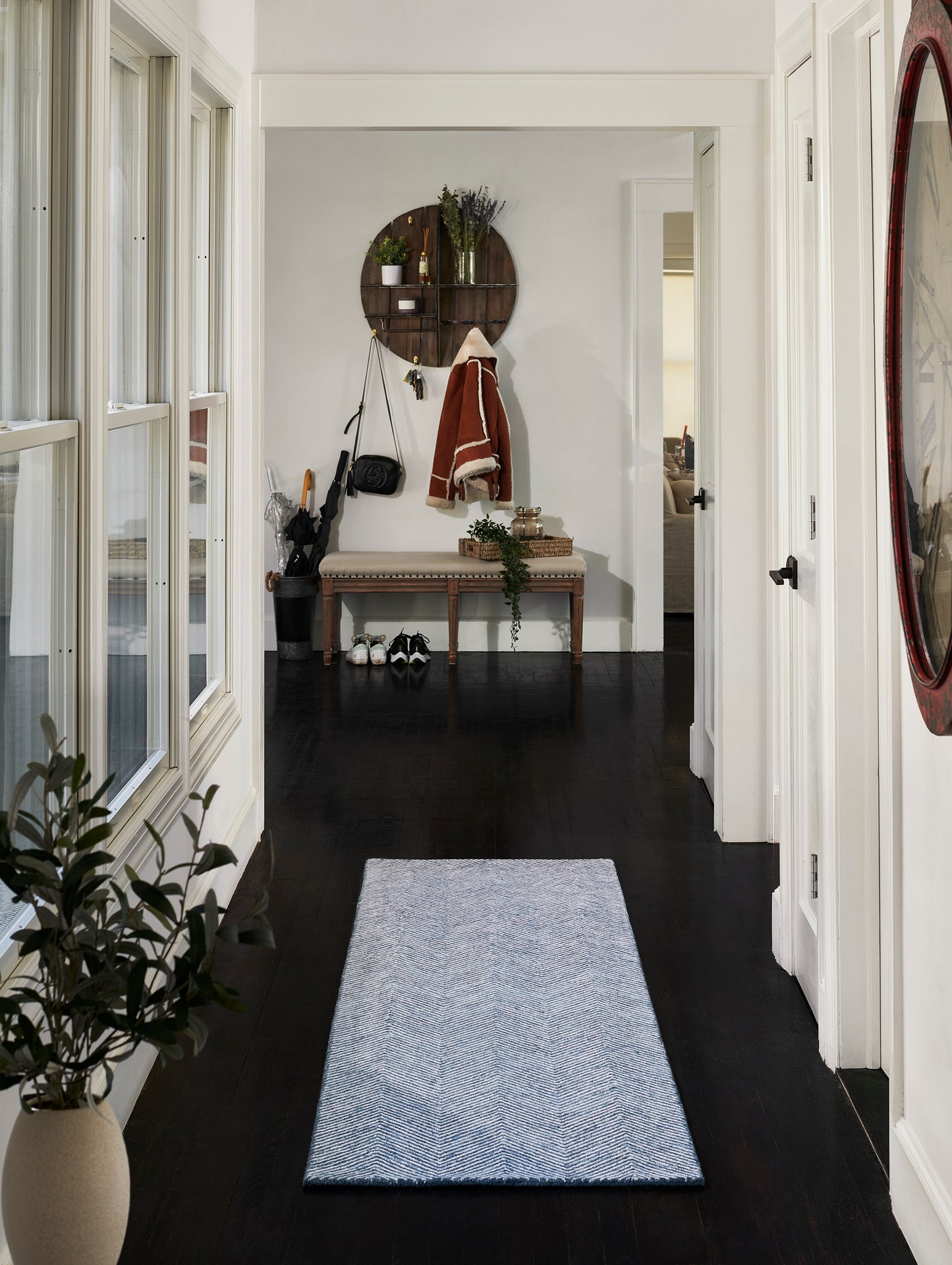 Narrow entry hall with dark wood floor, light blue chevron runner, white walls, round wood shelf with coats, beige bench, plants, and a red round mirror.