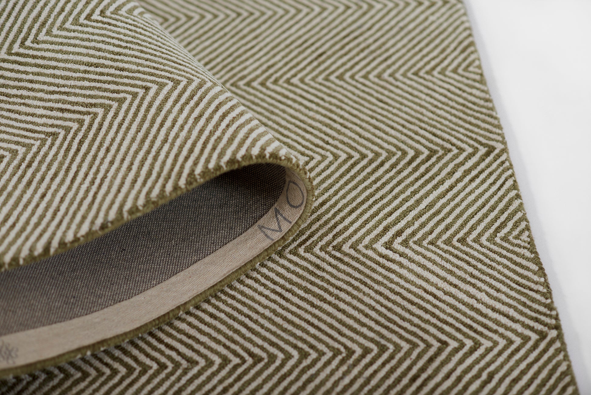 Olive green chevron woven rug with beige zigzag stripes, folded to reveal gray backing and tan edge.