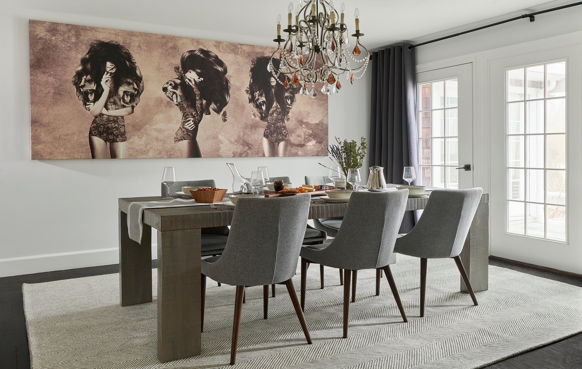 Grey upholstered dining chairs around a rustic wooden table beneath a crystal chandelier, sepia art, rug, glass doors.