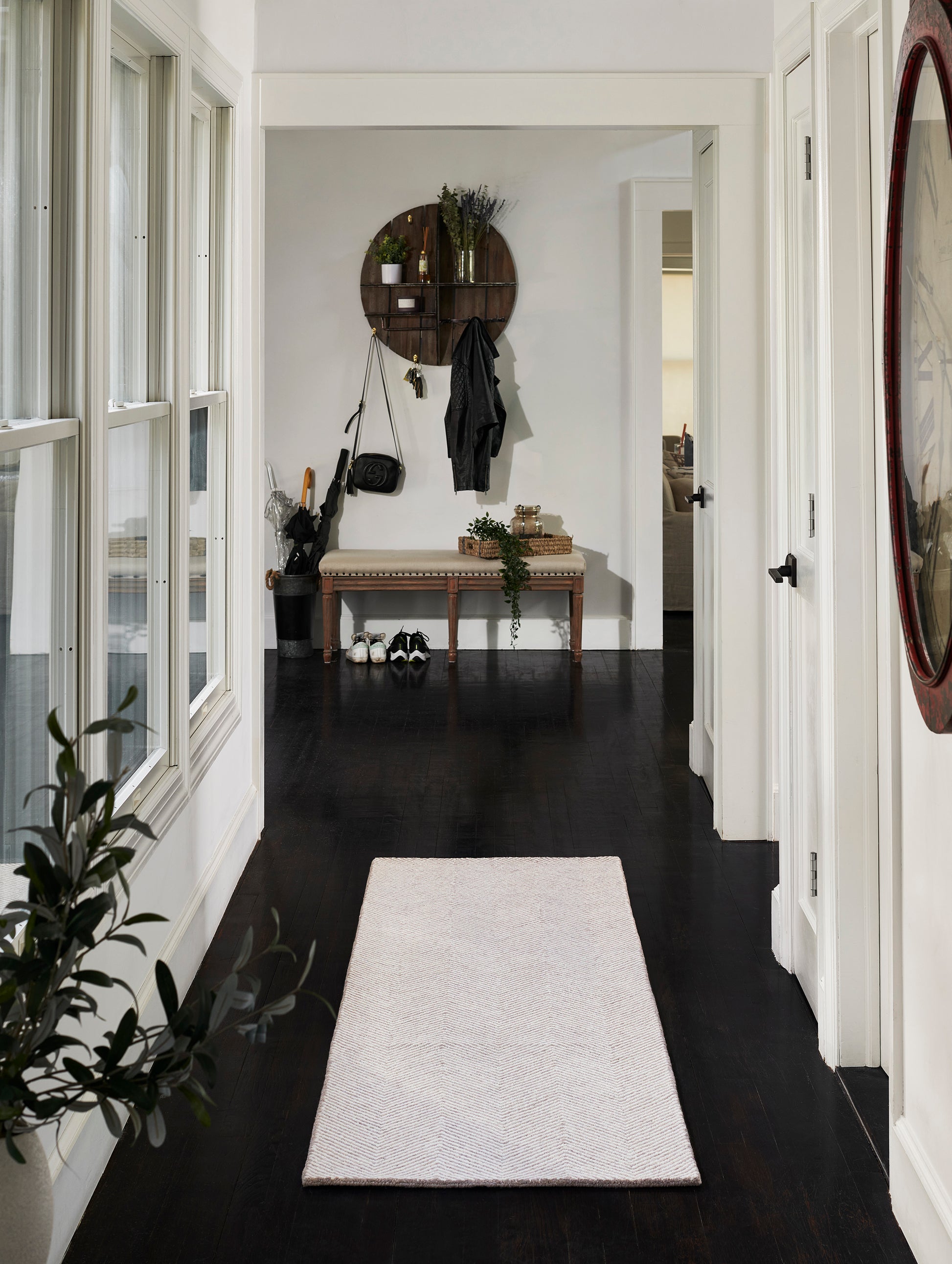 Taupe textured runner on dark wood hallway floor; bench under round wall shelf with coats, plants and a bag.