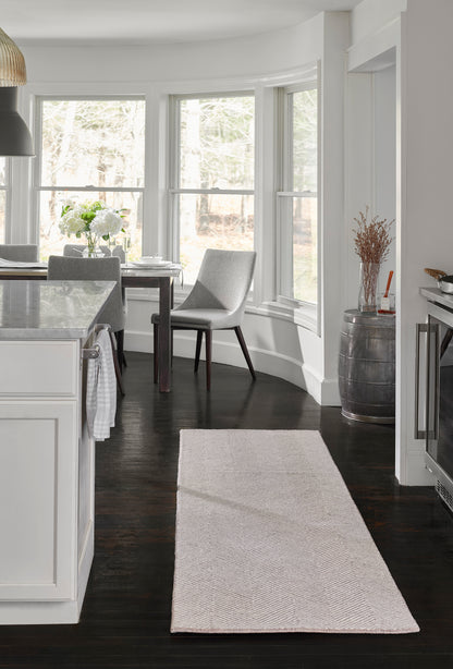 Taupe chevron-weave runner rug in a bright white kitchen/dining area with dark hardwood floor and gray chairs.