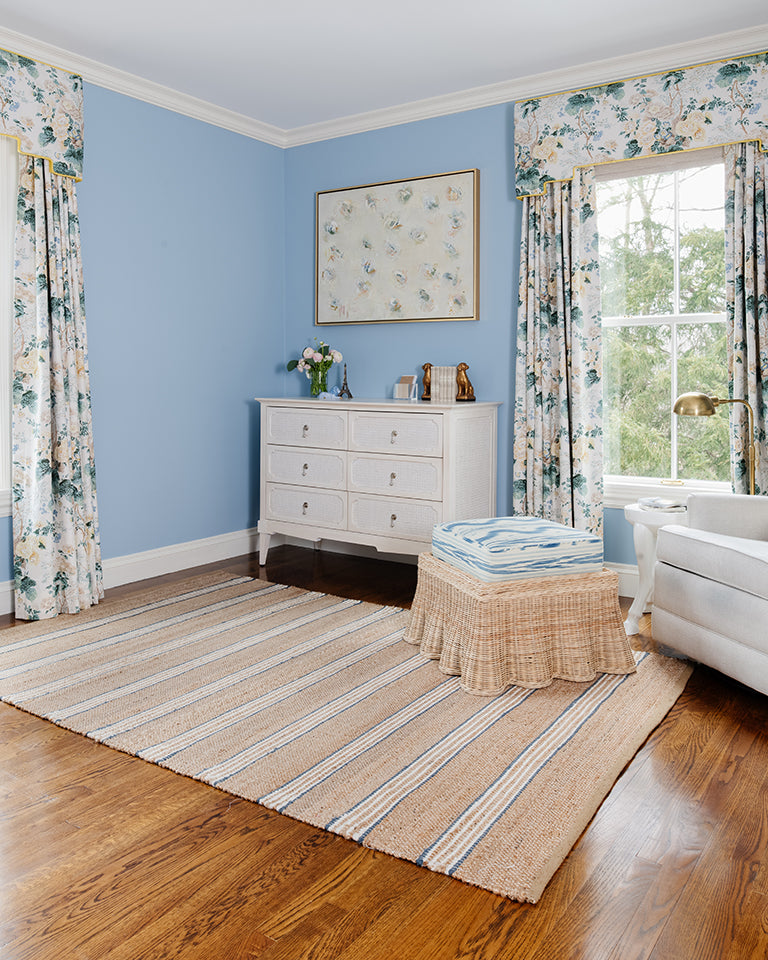 Blue-walled living room with white wicker chest, floral curtains, striped jute rug, and woven ottoman with blue throw.