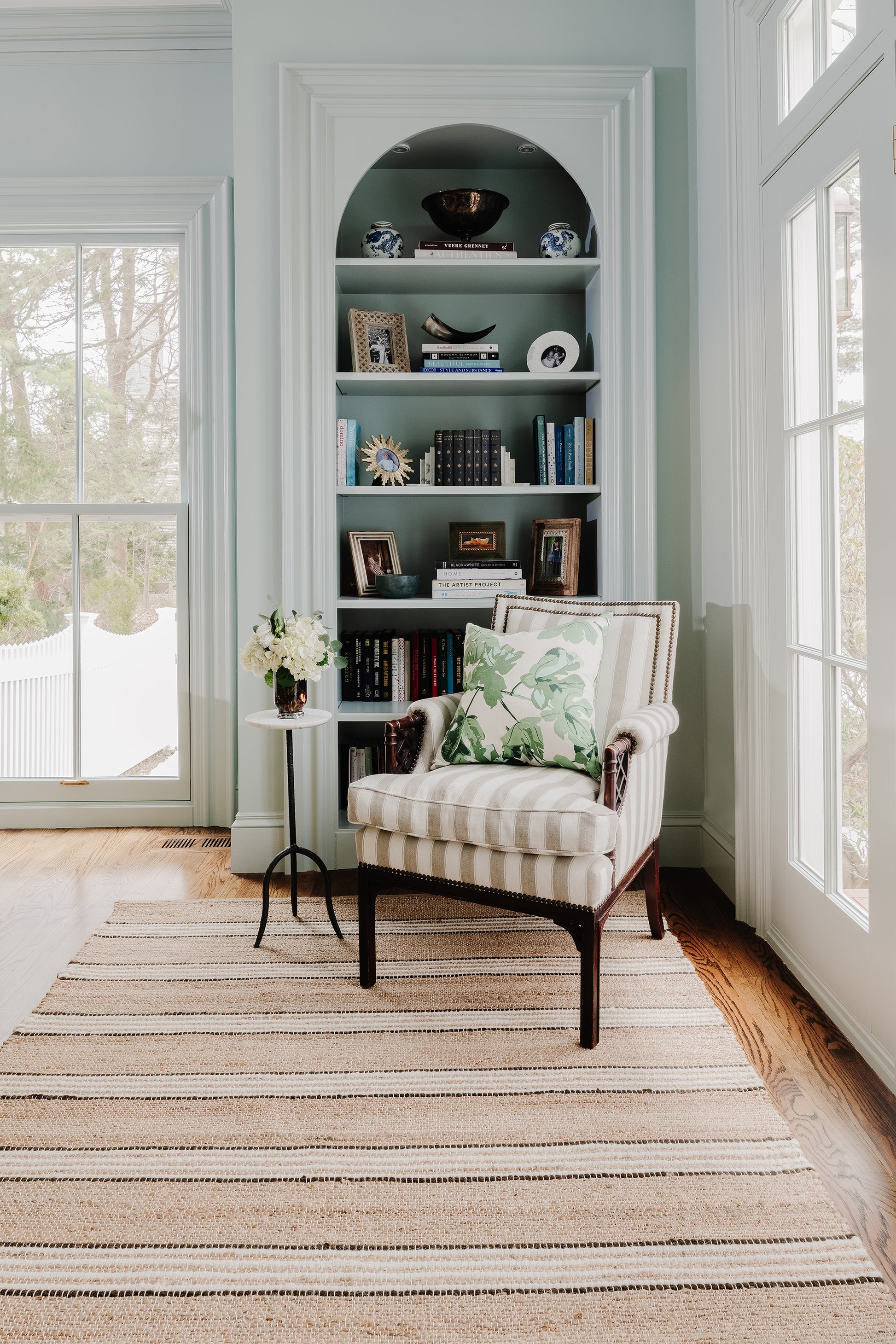 Arched built-in bookshelf in pale blue wall with books and decor, cream striped armchair and round side table by a window.