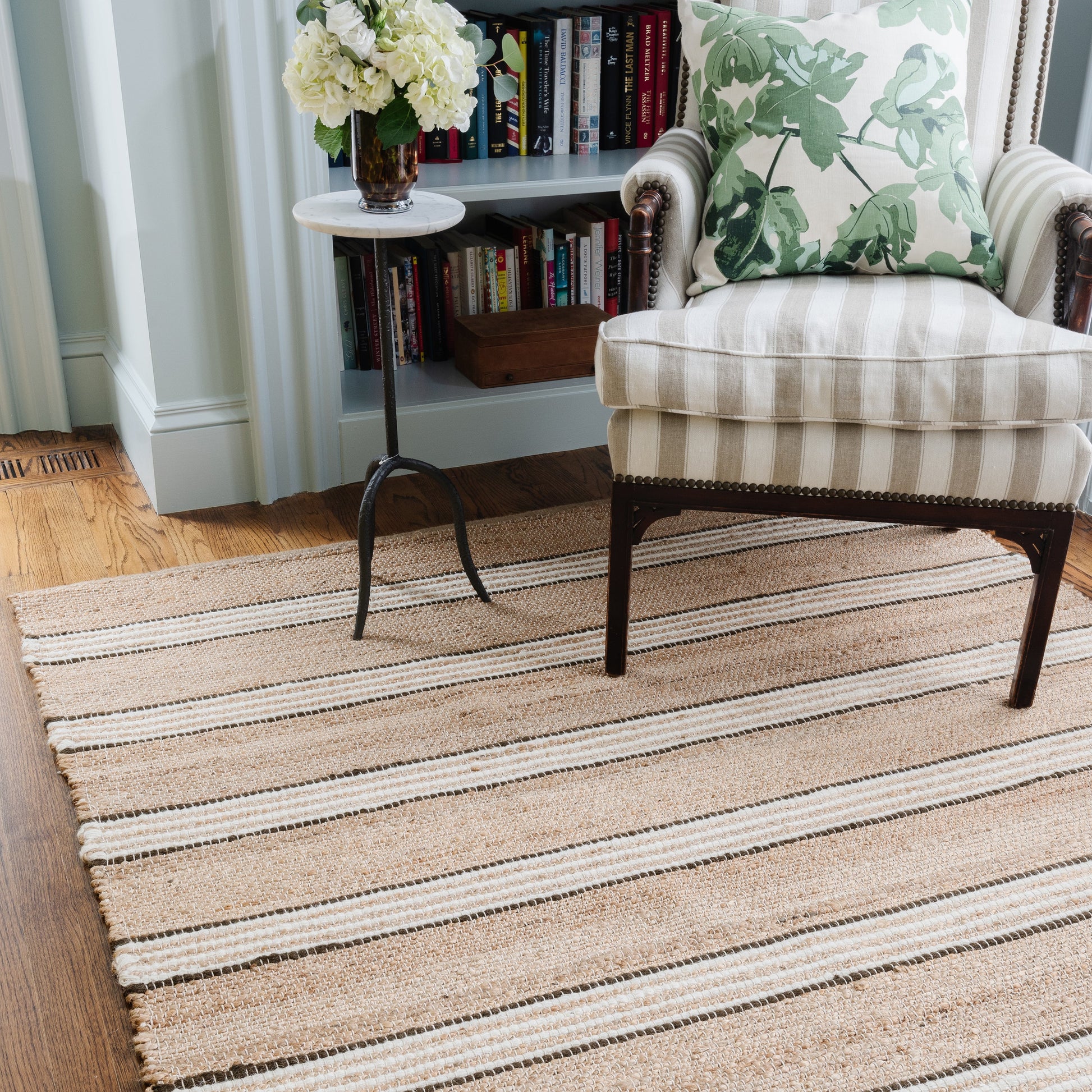 Beige striped jute rug beneath a striped armchair with a leaf-patterned cushion; round side table with flowers and a bookshelf behind.