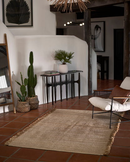 Natural beige jute area rug with fringed ends, boho living room scene with terracotta tile floor and a black console table.