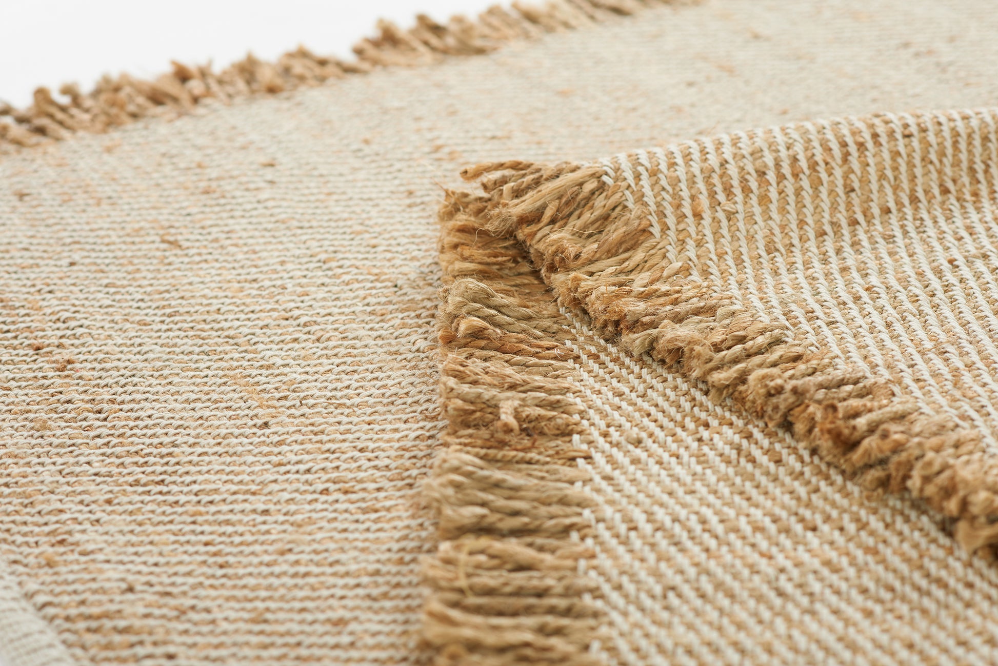 Natural beige jute rug with tan fringe tassels, folded to show thick woven texture.