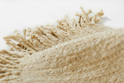 Beige woven jute rug with knotted fringe along the edge, natural fiber texture.
