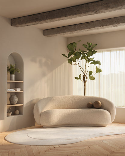 Cream bouclé rounded sofa on an oval rug in a bright neutral living room with exposed beams and a tall indoor plant.