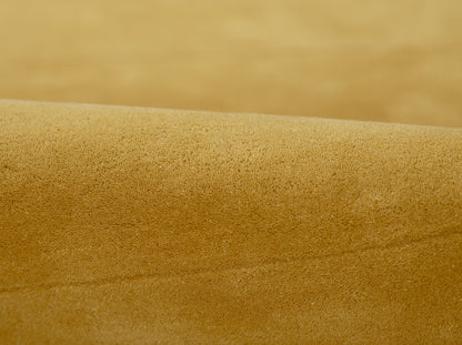 Golden tan suede fabric close-up with soft nap and gentle curved seam, warm desert-gold tone.