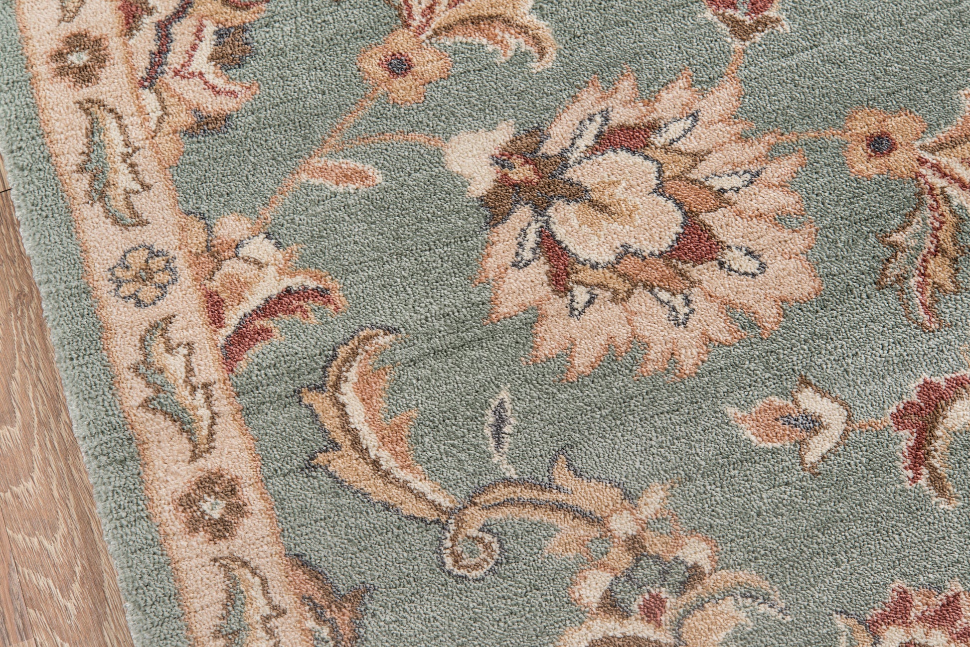 Teal floral rug with beige and cream flowers, pink accents, ornate border, wooden floor edge.