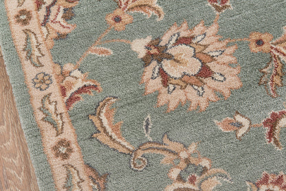 Teal floral rug with beige and cream flowers, pink accents, ornate border, wooden floor edge.