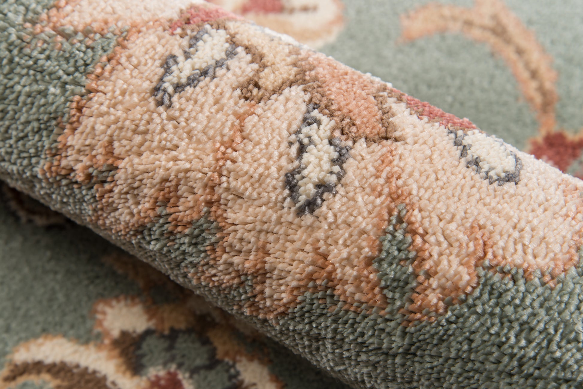 Close-up of a rolled oriental rug with green, peach, cream and brown tones in a plush loop pile.
