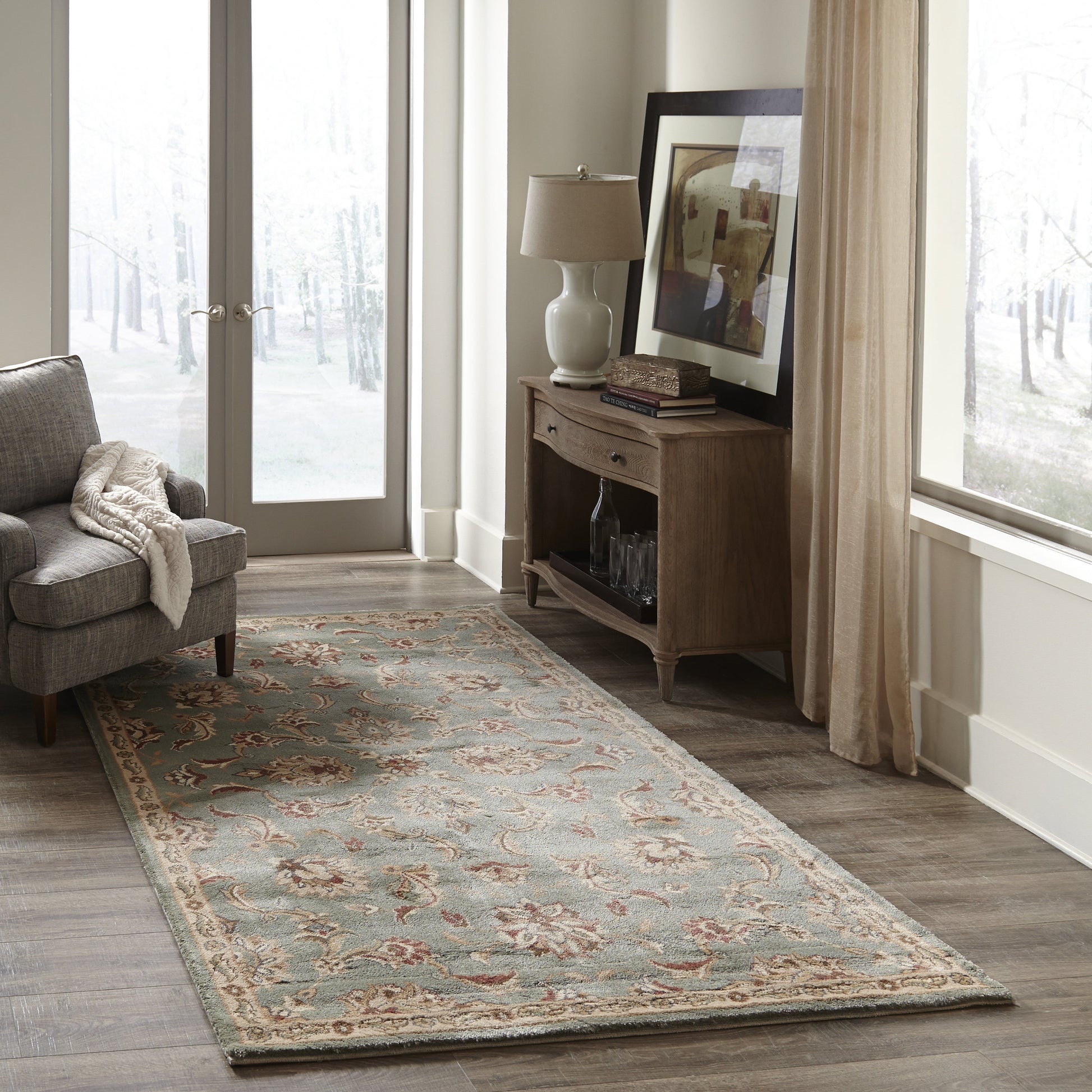 Traditional floral blue-beige area rug in a bright living room with a gray chair, wooden console, and large windows.