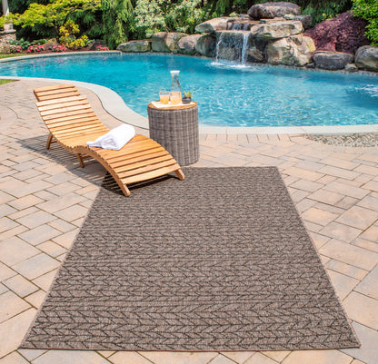 Tan & brown outdoor rug with geometric pattern beside a wooden pool lounge chair & wicker side table.