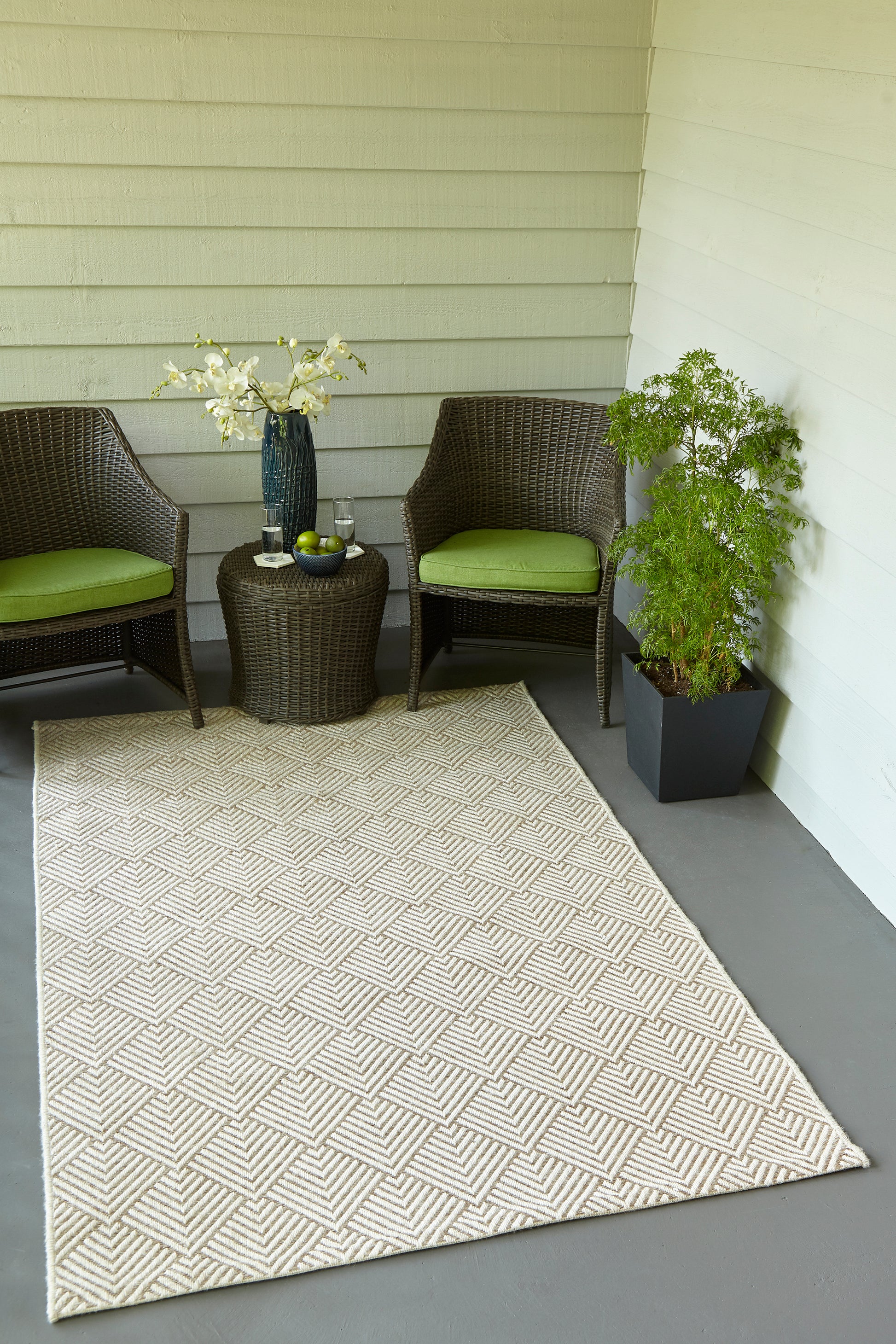 Outdoor wicker seating: brown chairs with green cushions, round table with white blossoms, beige geometric rug, potted plant.