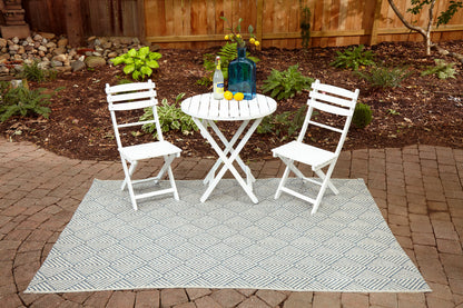 Outdoor patio scene with a white wooden bistro table and two folding chairs on a gray geometric rug over a brick patio; a blue vase with lemons.